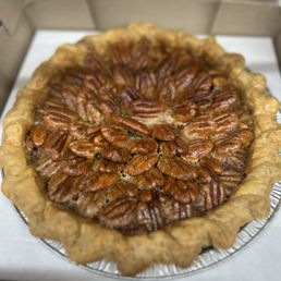 A pecan pie featuring a golden, flaky crust with a crimped edge. The filling is topped with a layer of glazed pecans arranged in a circular pattern. The pie is in a disposable aluminum pie pan, inside a cardboard box, ready for serving or delivery.