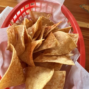 Pena's Place - Chips delicious and fresh - A red plastic basket contains a generous portion of crispy, golden-brown tortilla chips, lined with white paper. The basket is placed on a wooden table, with a drink in a clear glass cup visible in the background. The setting suggests a casual dining environment, likely at a Mexican restaurant or similar eatery. - New Mexican Cuisine Near Me - Ruidoso, New Mexico