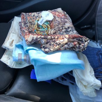 Savvy Consignment Boutique - A collection of clothing and accessories is arranged on a car seat, including a patterned scarf, a light blue shirt, and cream-colored pants. Gold and turquoise jewelry is placed on the scarf. The background shows the interior of a vehicle with black seats and door panels. - Thrift Stores Near Me - Brandon, Florida