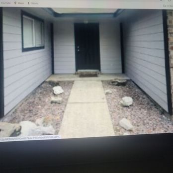 Palacios Construction Group - The image shows a front porch area with a concrete walkway leading to a black door, flanked by siding and a window, with rocks and gravel on either side. - General Contractors Near Me