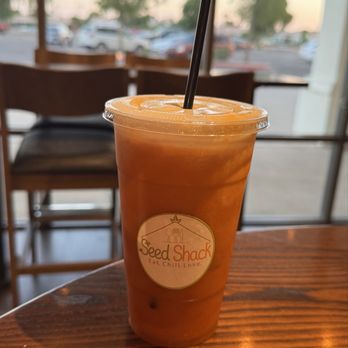 Seed Shack - Thai Tea - A clear plastic cup containing iced Thai tea with ice cubes and a black straw is positioned on a wooden table inside a cafe. The background shows large windows with a view of parked cars and trees outside, with soft lighting and a relaxed atmosphere. - Vegan Near Me - Gilbert, Arizona