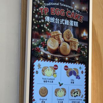 TP Tea - The digital menu display features images of traditional Taiwanese-style egg cakes with steam rising, set against a festive background with Christmas decorations. It highlights different flavors, including Yuzu Custard and Ube, marked as new, along with original and custard and boba options. The menu notes that each batch takes about five minutes to cook, and the display includes decorative holiday elements like snowmen and holly. - Bubble Tea Near Me - Flushing, New York