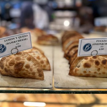 Cafe Baires - Two types of Argentinian pastries are shown on wooden boards in a bakery display. The left pastry is labeled 'Jamon y Queso (Ham & Cheese)' and the right is labeled 'Fugazzeta (Onion & Cheese)', each priced at $4.00. The background is blurred, highlighting the pastries and their labels. The image captures a bakery setting with a focus on traditional Argentinian baked goods. - Cafes Near Me - Hackensack, New Jersey