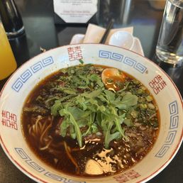 Beef Ramen - A bowl of ramen featuring a dark broth, noodles, and toppings such as fresh greens and herbs is shown on a restaurant table. The bowl has a decorative pattern with red and blue geometric designs. In the background, there are condiments, a glass of water, and a menu stand with specials listed. The setting appears to be a casual dining restaurant specializing in ramen.