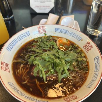Kaliubon - Beef Ramen - A bowl of ramen featuring a dark broth, noodles, and toppings such as fresh greens and herbs is shown on a restaurant table. The bowl has a decorative pattern with red and blue geometric designs. In the background, there are condiments, a glass of water, and a menu stand with specials listed. The setting appears to be a casual dining restaurant specializing in ramen. - Ramen Near Me - Wethersfield, Connecticut