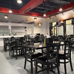 Interior of restaurant. A bunch of black tables and chairs. White bar in back. Orange beams make up the ceiling.