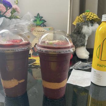 Deep Purpl - Two acai bowls with layered toppings, including fruit and granola, are positioned on a table. A yellow PATH flavored water bottle is placed beside them. The background features flowers, a plush toy wearing a hat, and some papers, indicating a casual setting. - Acai Bowls Near Me - Chicago, Illinois