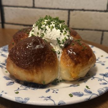The Dearborn - Parker House Rolls ($10) - A close-up of three baked potato skins on a white plate with blue floral patterns. Each potato skin is crispy and golden, topped with a dollop of sour cream and sprinkled with chopped chives. The plate is placed on a wooden surface, with a brick wall in the background, suggesting a casual dining setting. - New American Near Me - Chicago, Illinois