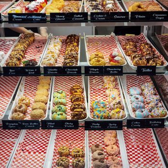 The Dapper Doughnut Cafe - Mini Donuts - A bakery display case showcases an assortment of decorated donuts and pastries, each labeled with flavor names, arranged on red and white checkered paper. - Coffee & Tea Near Me - Toronto, Ontario