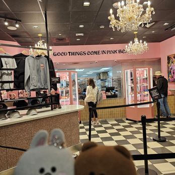 Voodoo Doughnut - Merchandise and chandeliers... look at that tiled floor! - A pink-themed retail store features chandeliers, clothing displays, and customers at the checkout counter, with plush toys in the foreground. - Donuts Near Me - Denver, Colorado