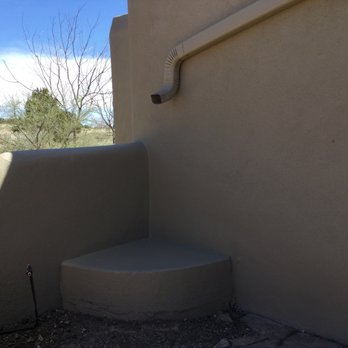 Always Construction Services - a corner of a building - The image shows a small outdoor corner with a beige stucco wall, a pipe attached to the wall, and a concrete bench built into the corner. The ground is uneven with some gravel and debris. In the background, there are trees and a partly cloudy sky. - Stucco Services Near Me - Santa Fe, New Mexico