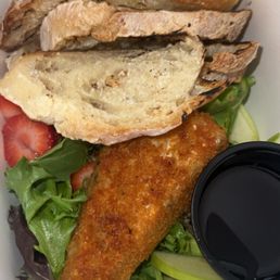 Fried Brie - A fried fish fillet with a golden-brown crust is placed on a bed of mixed greens, including lettuce and sliced cucumbers. Accompanying the fish are slices of bread, some with a spread or topping, and a few strawberry slices. A small black container filled with dark sauce is also included. The meal is presented in a white takeout box.
