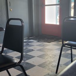 Front door loxated 1 vlock northwest of marked lication. - The image shows the interior of a small dining area with a black and white checkered tile floor. There are black chairs positioned around the space, and a door with a window that has signs and notices attached. The door appears to lead outside, with sunlight visible through the window. The walls are painted a dark color, and there is a small red sign on a gray door to the left. The setting suggests a casual eatery or cafe.