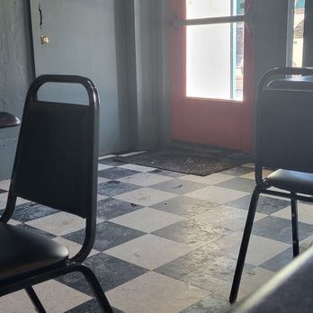 Big Head Burger - Front door loxated 1 vlock northwest of marked lication. - The image shows the interior of a small dining area with a black and white checkered tile floor. There are black chairs positioned around the space, and a door with a window that has signs and notices attached. The door appears to lead outside, with sunlight visible through the window. The walls are painted a dark color, and there is a small red sign on a gray door to the left. The setting suggests a casual eatery or cafe. - Burgers Near Me - Waterloo, Iowa