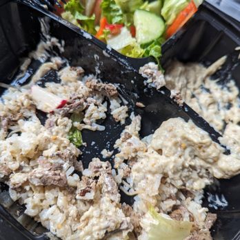 Pita Thyme - Not sure what I just ate. Can't even finish it - The image shows a black plastic takeout container with two compartments. One compartment contains a salad with chopped cucumbers, lettuce, and tomato pieces. The other compartment has a creamy dish with rice, ground meat, and possibly cheese or sauce, with some remnants of food and a fork resting on the container. The food appears to be from a restaurant offering Mediterranean or Middle Eastern cuisine. - Lebanese Near Me - Boston, Massachusetts