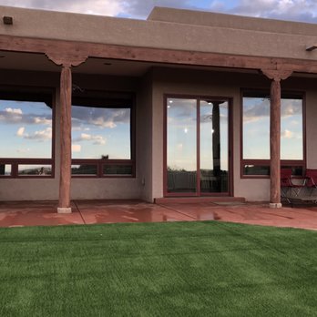 AMG Landscape - a view of a house with a patio and lawn - The image shows the exterior of a building with large windows, wooden columns, and a patio area with chairs and artificial grass. - Irrigation Near Me