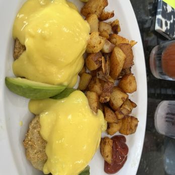 Maria's Cafe - A white oval plate holds two eggs Benedict, each topped with a generous amount of hollandaise sauce. The dish is garnished with slices of avocado and served alongside a portion of diced, fried potatoes. The presentation is simple, with ketchup on the side. The background shows a dark countertop with some objects, including a glass of drink and a pack of cigarettes. - Breakfast & Brunch Near Me - Stuart, Florida