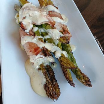Provision - Asparagus - The image shows three asparagus spears wrapped in bacon, topped with a creamy white sauce, and placed on a white rectangular plate. The asparagus appears grilled with some charring, and the bacon is cooked to a crisp. The dish is garnished with a generous amount of the sauce, which covers parts of the asparagus and bacon. The plate is set on a wooden table, and the photo includes text labels indicating the dish as 'Bacon Wrapped Asparagus'. - New American Near Me - Indianapolis, Indiana