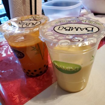 BAMBU - Drinks ordered with no ice and one is barely half full. Skimping out on drinks. - Two cups of bubble tea with sealed plastic lids are positioned on a table at Bambu restaurant. One drink contains dark tapioca pearls, and the other appears to be a creamy, light-colored beverage. The cups have Bambu branding and are placed on a red and white tablecloth. In the background, there are various items including a paper towel roll, a plastic container, and a bowl with foil-wrapped items, indicating a casual dining setting. - Bubble Tea Near Me - West Allis, Wisconsin