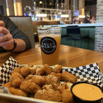 Crust Brewing - Spicy cheese curds - A tray of fried cheese curds topped with grated cheese and served with a side of dipping sauce is placed on a checkered paper liner on a wooden table. Behind the tray, there is a glass of beer with the Crust Brewing logo. The background shows the interior of Crust Brewing, with blurred patrons, brick walls, and modern lighting fixtures, indicating a casual dining environment. - Breweries Near Me - Rosemont, Illinois
