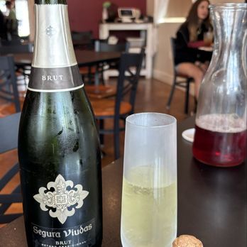 Stem & Stone - Cranberry mimosa coming up - The image shows a restaurant table with a bottle of Segura Viudas Brut Cava, a tall flute glass filled with sparkling wine, and a cork with a wire cage. In the background, there are chairs, a woman sitting at another table, and restaurant interior elements such as curtains and a wall with a picture. The setting appears casual and well-lit. - New American Near Me - Bastrop, Texas