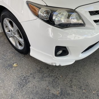 NTC Auto Body - Side picture - The image shows the front right side of a white sedan with noticeable scratches and scuff marks on the lower part of the front bumper. The car is parked on an asphalt surface with some small debris. The headlight and wheel are visible, and the damage is concentrated near the corner of the bumper. - Body Shops Near Me - Fishkill, New York