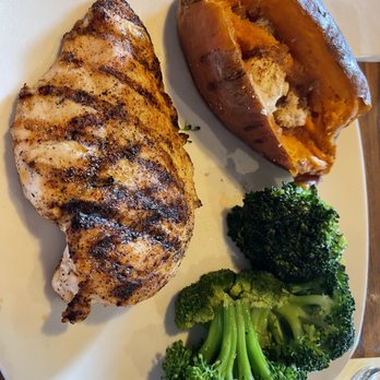 Outback Steakhouse - Chicken on the Barbie with loaded sweet potato and steamed broccoli... delish!! - The image shows a meal with a grilled chicken breast with visible grill marks, a baked sweet potato with some of its flesh exposed, and a portion of steamed broccoli florets. The food is arranged on a white, oval-shaped plate, placed on a wooden table. The meal appears to be a balanced, healthy dish with protein, vegetables, and a carbohydrate source. - Steakhouses Near Me - Dover, Delaware