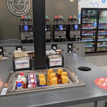 RaceTrac - RaceTrac, Ormond Beach - The image depicts a coffee station in a convenience store, featuring several beverage dispensers on a counter, with a refrigerated section filled with bottled and canned drinks in the background. The station includes a napkin dispenser and a sign instructing to use a napkin or not. The area is clean and organized, with a tiled floor and store branding visible on the wall. - Gas Stations Near Me - Ormond Beach, Florida