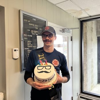 Roberto's Bakery - 21st Birthday - A man wearing glasses, a cap, and a dark shirt stands indoors holding a birthday cake with candles, celebrating a 21st birthday. - Bakeries Near Me - Taylor, Michigan