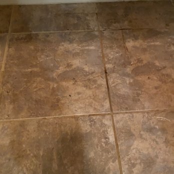 Texas Cleaning Services - Pantry floor was not cleaned - The image depicts a section of a tiled floor in a pantry or storage area. The tiles are brown with a marbled pattern, and there are visible stains, dirt, and small debris scattered across the surface. The background includes a white baseboard or wall trim at the top of the image. The overall appearance suggests the floor has not been recently cleaned. - Home Cleaning Near Me - Austin, Texas