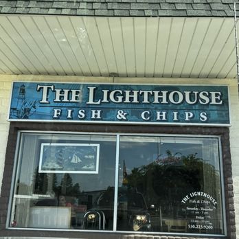 The Lighthouse - Front of Building - The image shows the exterior of The Lighthouse Fish & Chips restaurant, with a prominent blue sign displaying the business name and a lighthouse graphic. The sign reads 'The Lighthouse Fish & Chips.' The storefront has a large window with the restaurant's hours and contact number, and the reflection of the parking lot and trees is visible in the glass. The building has a shingled roof and a beige exterior wall. - Seafood Near Me - Redding, California
