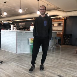 Tempest with the buddy! - A man with glasses and a beard stands inside a coffee shop, wearing a black hoodie with a colorful logo on the chest and black pants. He is smiling and has his hands in his pockets. The coffee shop has a counter with a sign that reads 'TEMPEST COFFEE COLLECTIVE,' and there are hanging light bulbs from the ceiling. The interior features a light-colored wooden floor and a dark wall with shelves and a small table in the background.