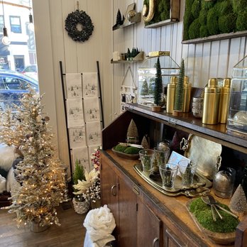 Bench492 - November 2021. - The image shows a shop interior decorated for the holiday season, with a small, illuminated Christmas tree on the left side, surrounded by ornaments and decorative items. A wooden cabinet holds glassware, trays, and small decorative trees, with additional holiday decor on top. The wall features shelves with ornaments, greenery, and decorative objects, including gold containers and moss-covered letters. The space has a warm, inviting atmosphere with natural light coming through a window. - Home Decor Near Me - Glen Ellyn, Illinois