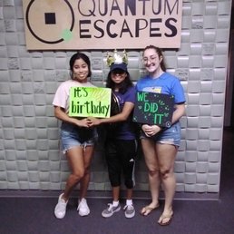 This is when we finished the room - Three women stand together in front of a wall with a large sign reading 'Quantum Escapes,' an escape room business. They hold signs, one reading 'It's my birthday' and another 'We did it.' The woman in the middle wears a crown and a cap, indicating a celebration. The women are smiling and dressed casually, suggesting they have completed an escape room challenge at the venue.
