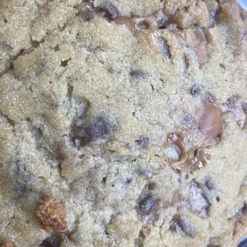 Insomnia Cookies - Salted Caramel Deluxe Cookie - A close-up image of a salted caramel deluxe cookie showing a golden-brown surface with visible chocolate chips and caramel chunks. The cookie has a crumbly texture with some areas appearing slightly cracked, and the caramel and chocolate pieces are embedded throughout. The image emphasizes the cookie's rich ingredients and baked appearance, likely taken at a bakery or food establishment. - Desserts Near Me - Albuquerque, New Mexico