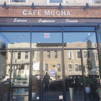 Cafe Mocha - The image shows the storefront of Cafe Mocha in Queens, NY, with large glass windows reflecting nearby buildings and parked cars. The facade includes a brick section with the cafe's name, 'CAFE MOCHA,' displayed at the top, and a black awning with white lettering advertising espresso, cappuccino, and desserts. Potted plants are positioned on either side of the entrance, and the interior is partially visible through the windows. - Cafes Near Me - Queens, New York