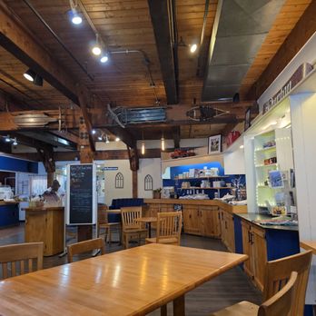 Three Mountain Cafe - Inside - The image shows the interior of a cafe with wooden furniture, including tables and chairs, and a wooden ceiling with exposed beams. The decor includes blue and white walls, with shelves displaying various items. The counter area is visible on the left, with a staff member working behind it. The space is well-lit with ceiling spotlights, creating a warm and inviting atmosphere. - Sandwiches Near Me - Waitsfield, Vermont