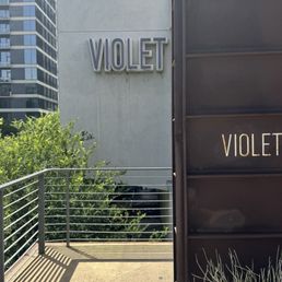 Violet signage is displayed on a building exterior with a cityscape background, including trees and modern high-rise buildings.