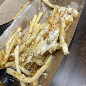 SubKO - Garlic truffle fries - French fries with melted cheese and seasoning are placed in a rectangular cardboard container. The fries are thin-cut and some are coated with cheese, which appears melted and slightly gooey. The container is on a dark wooden surface, and a black plastic fork is partially visible in the corner. The fries are seasoned with black pepper and possibly other spices, and some cheese has spilled over the sides of the fries. - Sandwiches Near Me - Lisle, Illinois