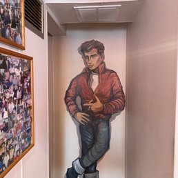 A mural of a young man in a red jacket and jeans is painted on a wall in an indoor setting, with photos on the adjacent wall.