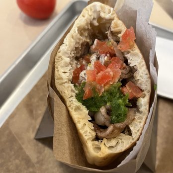Miznon - Steak pita - A close-up of a steak pita sandwich filled with chopped tomatoes, greens, and meat, wrapped in paper, with a tomato in the background. - Middle Eastern Near Me - Toronto, Ontario