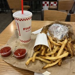 Meal - The image shows a meal served at Five Guys, featuring a large portion of French fries in a paper cup, a burger wrapped in aluminum foil, and two small containers of ketchup. A large soft drink in a branded cup with a red straw is also present. The meal is placed on a wooden table inside a casual dining area with wooden chairs and tables, and a red-and-white checkered wall in the background.