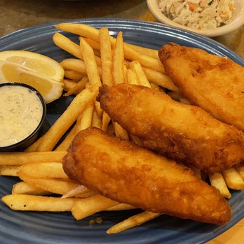 O'Niell's - Fish and chips - A blue ceramic plate holds three pieces of fried fish fillets, French fries, lemon wedges, and a small container of tartar sauce. The plate is garnished with a leaf of lettuce. In the background, there is a bowl of coleslaw and a glass of beverage, indicating a meal at a restaurant. The dish appears to be a classic fish and chips presentation. - Irish Near Me - Albuquerque, New Mexico