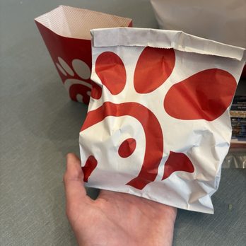 Chick-fil-A - Chick Fil a Chick-fil-A- Sandwich - A hand is holding a crumpled Chick-fil-A paper bag with a red and white design featuring the Chick-fil-A logo. In the background, there is a Chick-fil-A cup and a white plastic bag on a gray surface, with some indistinct objects at the top of the image. The setting appears to be a table or counter, possibly in a restaurant or break area. - Fast Food Near Me - Stuart, Florida