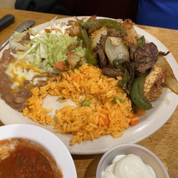 Three Amigos - Chicken and Steak Fajitas - A large plate features Mexican dishes including rice, salad with guacamole, beef with onions and peppers, and a side of salsa and sour cream. - Mexican Near Me - Stoneham, Massachusetts