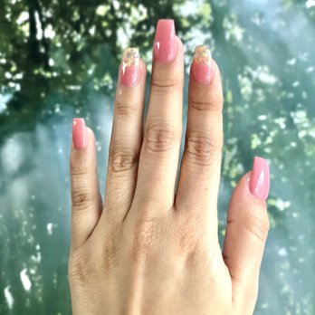 New York Nails 2 - Short coffin nails done by Giah! - A hand with well-maintained, glossy pink nails is displayed prominently in the foreground. The nails are decorated with gold accents on the tips, creating a stylish manicure. The background is an out-of-focus outdoor scene with trees and sunlight filtering through the leaves, providing a natural setting for the image. - Nail Salons Near Me - Pacifica, California