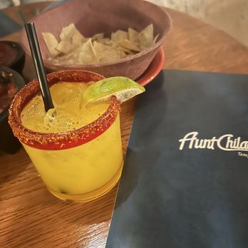 Aunt Chilada's - Skinny Mango Margarita - A yellow margarita with a salted rim, lime wedge, and straw is placed on a wooden table at Aunt Chilada's in Tempe, Arizona, with side dishes in the background. - Mexican Near Me - Tempe, Arizona