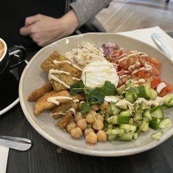 Cafe Demi - Jerusalem chop salad with the gluten croutons and a scoop of tuna. - A plate contains a variety of fresh vegetables and toppings, including chickpeas, chopped cucumbers, diced tomatoes, and a poached egg in the center. The dish is garnished with herbs and drizzled with a creamy sauce. To the side, there is a cup of coffee with latte art, served in a black cup with a saucer. The setting appears to be a cafe with a dark wooden table and utensils placed nearby. - Coffee & Tea Near Me - Teaneck, New Jersey