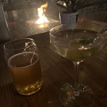 3128 Social House - Our view - The image shows a wooden table with a glass of beer and a cocktail in a martini glass, both positioned near a window. A small potted plant with a label marked '31' is also on the table. Outside the window, a fire is burning, providing warm lighting to the scene. The setting appears to be indoors, possibly in a restaurant or bar, with a cozy ambiance created by the fire and dim lighting. - Gastropubs Near Me - Albuquerque, New Mexico