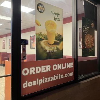 Desi Pizza Bite - Order online - The storefront window shows two posters: one promoting Mango Lassi with an image of a yellow drink and the text 'Refresh Your Senses,' and another advertising pizza with the phrase 'Greatest of Flavors.' Below, a large sign encourages customers to order online at desipizzabite.com. The interior of the restaurant is visible through the glass, with seating and wall decorations. - Pizza Near Me - Tracy, California